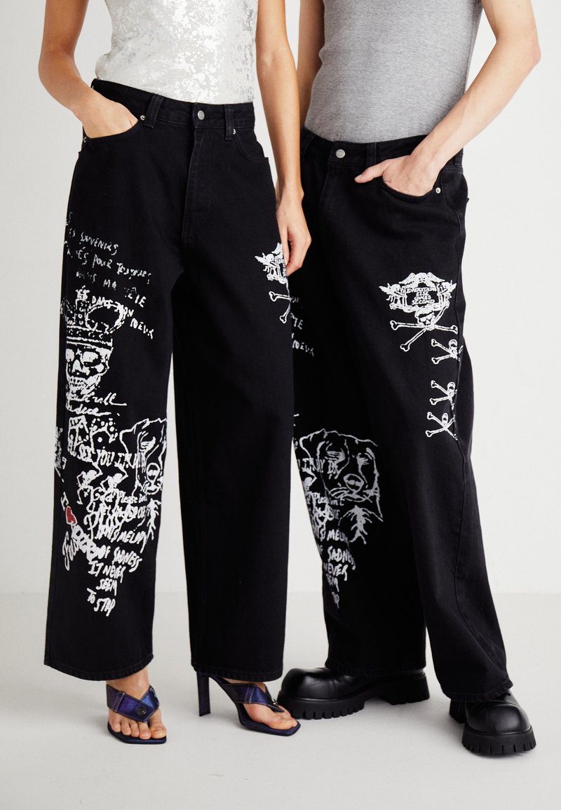 Jeans en denim noir à large jambe présentant des impressions graphiques blanches de crânes et de chiens, avec deux poches avant et une coupe décontractée.