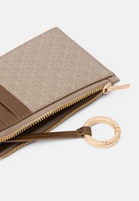 Portefeuille beige avec motif géométrique, accents marron, fermeture éclair et anneau dorés. Comprend des emplacements pour cartes et un compartiment zippé. Texture lisse en synthétique.