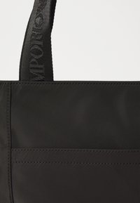 Borsa tote in nylon nero con una texture liscia, tasca frontale e tracolla intrecciata con dettagli di branding. Forma semplice e rettangolare.