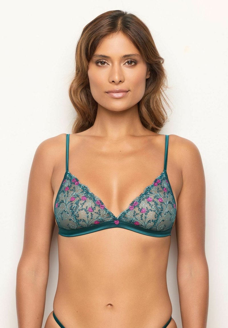 Donna che indossa un reggiseno in pizzo trasparente turchese con ricami floreali rosa, in piedi contro uno sfondo chiaro e semplice.