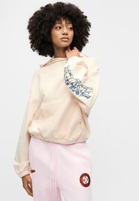 Ροζ φούτερ με κουκούλα και κεντημένο μπλε κείμενο στο μανίκι, σετ με ροζ παντελόνι φόρμας που φέρει κυκλικό κόκκινο έμβλημα.