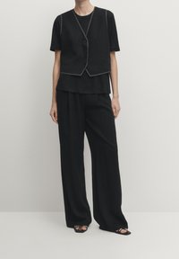 Schwarzes Outfit mit einem kurzärmeligen Oberteil mit geschichteter Gestaltung, einem taillierten Weste mit Knopfdetails und weit geschnittenen Hosen aus einem strukturierten Stoff.