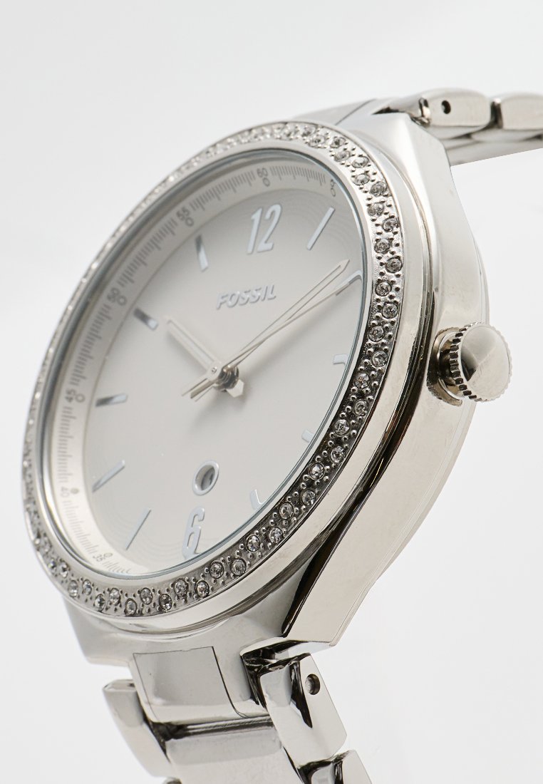 Montre-bracelet en argent avec un cadran rond, une lunette ornée de cristaux, un cadran blanc et un bracelet en métal avec des maillons polis.