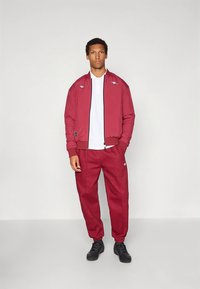 Fato de treino bordô composto por um casaco tipo bomber com fecho éclair e calças combinando, com um logótipo branco, contrastando com uma camisola pólo branca por baixo.