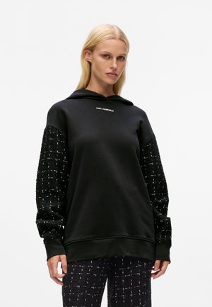Sudadera negra con mangas negras texturizadas que presentan un patrón de cuadros blancos. Tejido texturizado y detalle del logo cerca del escote. Diseño casual.