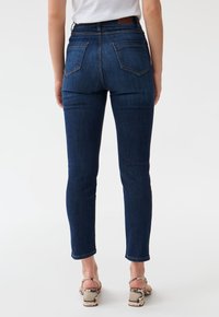 Dark blue denim skinny jeans with a high waist, featuring two back pockets and a subtle fade. Styled with heeled sandals.