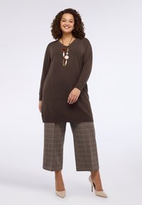 Tunika marrone a maniche lunghe con scollo a V. Abbinata a pantaloni a gamba larga grigi a quadri. Accessori includono una collana con ciondolo multicolore e tacchi color nudo.