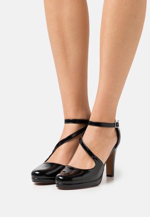 Chaussures à talons hauts en cuir verni noir avec un bout arrondi, une lanière à la cheville et un talon de 12,7 cm. Le design présente une finition lisse et brillante.