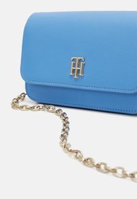 Malá modrá strukturovaná kožená kabelka se zlatým řetízkovým popruhem a zlatým logem "TH" na předním klopě na bílém pozadí.