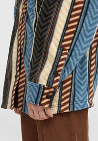 Fleece jacket with herringbone pattern in blue, beige, and brown. Features black buttons and a side pocket. Corduroy pants visible below.