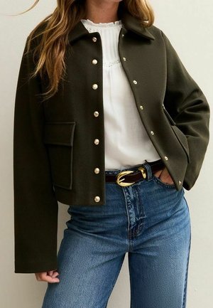 Kobieta ubrana w ciemnozieloną kurtkę zapinaną na guziki, na białej bluzce wsuniętej w wysokowodne niebieskie dżinsy z brązowym paskiem.