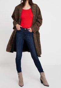 Abrigo a cuadros marrón y naranja, top rojo ajustado, jeans skinny azul oscuro y botines beige con tacón bloque. Conjunto casual en capas.