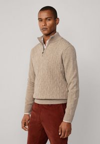Beige Kabelstrickpullover mit hohem Kragen und halbem Reißverschluss. Gerippte Bündchen und Saum, getragen über einem karierten Hemd.