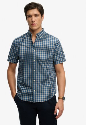 Junger Mann trägt ein blau-grünes kariertes kurzärmeliges Hemd, dunkle Hose und eine Armbanduhr, steht mit teilweise in den Taschen steckenden Händen.