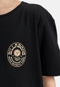 Zwarte T-shirt met een tan Billabong-logo waarop een zon, golven en de tekst "Boardshort Co. Sinds Drieëntwintig" staat.