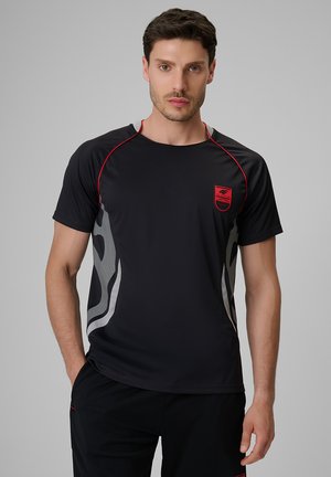 Mężczyzna ubrany w czarną sportową koszulkę z czerwonymi lamówkami i logo, stojący z jedną ręką w kieszeni na jednolitym szarym tle.
