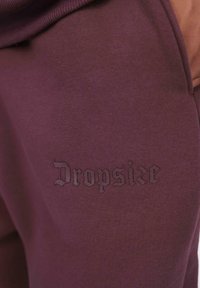 Nahaufnahme von kastanienbraunem Stoff mit "Dropsike" in passendem dunklem Faden auf der Kleidung gestickt.