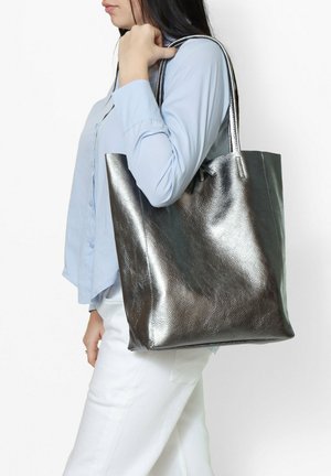 Metalen zilveren tote bag met een zachte textuur, voorzien van twee lange hengsels en een open bovenkant, gedragen op de schouder.