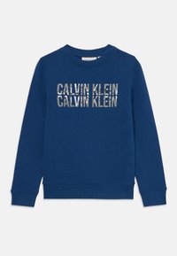 Blågenser laget av bomull, med rund hals, ribbestrikkede mansjetter og kant. Framsiden viser "CALVIN KLEIN" i metallisk trykk.
