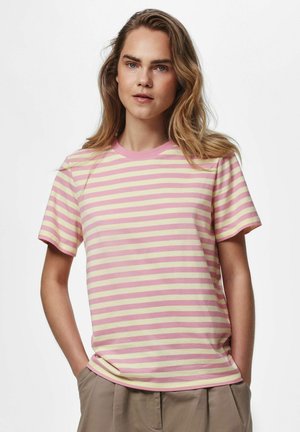 Vrouw met lang blond haar draagt een roze met crème horizontaal gestreept T-shirt en beige broek, staat met handen in de zakken tegen een effen achtergrond.