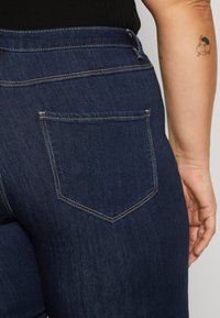 Jeans de mezclilla oscura con un corte ajustado, que cuentan con un bolsillo trasero con una solapa puntiaguda y detalles de costura. Tejido texturizado, peso medio.