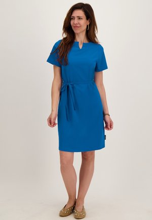 Vrouw draagt een knielange blauwe jurk met korte mouwen en een taillekoord, staat en kijkt iets schuin opzij tegen een effen achtergrond.