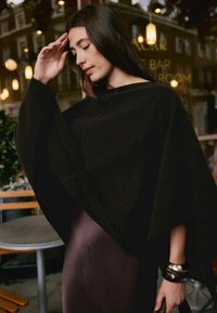 Black knit poncho with loose sleeves, draping design, paired with a dark purple smooth dress and layered bracelets on the wrist.