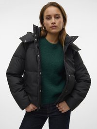 Svart puffjakke med høy krage, trykknapper og en blank finish, lagd over en grønn teksturert genser mot en ensfarget bakgrunn.