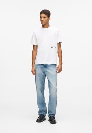 T-shirt en coton blanc avec un col rond, arborant un petit logo noir sur le côté gauche, associé à un jean en denim bleu clair et des chaussures noires.