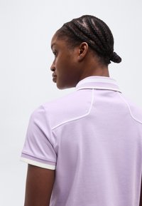 Άτομο με πλεξούδες μαλλιών δεμένες κότσο, που φοράει ανοιχτό μωβ κοντομάνικο πουκάμισο με άσπρο περιθώριο, στραμμένο προς τα αριστερά μπροστά σε απλό φόντο.