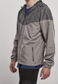 Man som bär en tvåfärgad grå huva jacka med elastiska ärmar och fickor med dragkedja över en marinblå skjorta och svarta byxor.