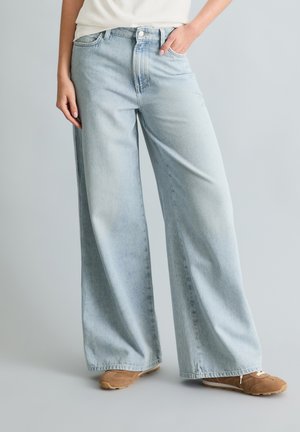 Person trägt hellblaue, weit geschnittene Jeans und braune Turnschuhe, steht vor einem schlichten hellgrauen Hintergrund.