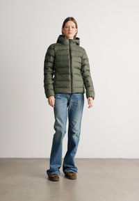 Chaqueta acolchada verde oliva con capucha, con cierre de cremallera en la parte delantera y diseño de acolchado horizontal. Llevada con jeans de campana azules y zapatos de estampado de leopardo.