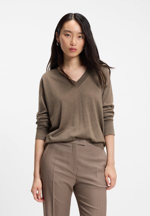 Jeune femme aux longs cheveux foncés portant un pull marron à col en V et un pantalon taupe sur fond blanc.
