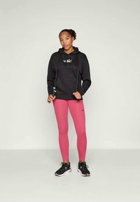 Sweatshirt preto com capuz e logotipo, leggings cor-de-rosa e sapatilhas de desporto pretas. De pé com uma corda de saltar, fundo neutro.