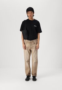 Zwarte t-shirt met subtiele logo, lichtbeige recht gesneden jeans, zwarte schoenen met witte accenten, en een kleine zwarte crossbody tas.