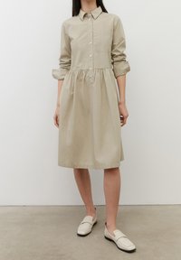 Abito beige con colletto tondo, vita arricciata, maniche lunghe e tasca sul petto, abbinato a scarpe slip-on di colore chiaro.