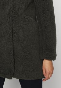 Abrigo de forro polar en gris oscuro con una superficie texturizada, que presenta un corte relajado, bolsillos laterales y mangas de longitud media.