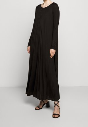 Femme portant une longue robe noire plissée à manches longues et des sandales noires à talons avec brides, debout devant un fond clair uni.
