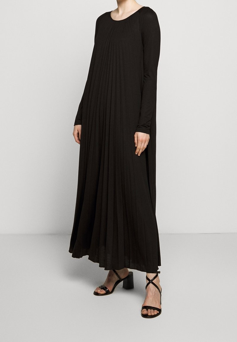 Femme portant une longue robe noire plissée à manches longues et des sandales noires à talons avec brides, debout devant un fond clair uni.