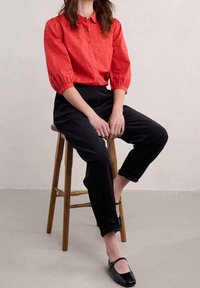 Camicia rossa con bottoni, decorata con piccoli punti bianchi e maniche a sbuffo. Pantaloni in velluto a coste neri e scarpe Mary Jane nere. Modella seduta su uno sgabello di legno.