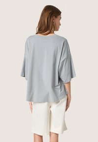 Grå oversized T-shirt med korte ærmer, der har små glitrende accenter. Sammen med beige knælange shorts. Glat bomuldsstruktur.