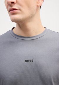 Šedé tričko s krátkym rukávom a okrúhlym výstrihom, na hrudi s nenápadným čiernym logom "BOSS". Hladká textúra, neformálny dizajn.