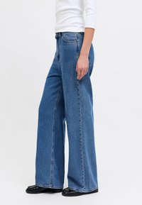 Person iført løstsiddende blå jeans, hvid langærmet top og sorte sko, stående mod en ensfarvet lys baggrund.