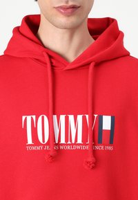 Felpa rossa con cappuccio e coulisse, con il testo "TOMMY" in grandi lettere bianche e accenti del logo. Tessuto morbido.