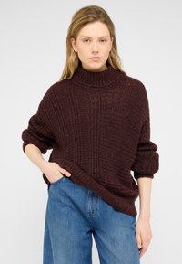 Bordo apykaklė megzta suknelė su aukštu kaklu, tekstūruotu raštu ir per dideliu siluetu. Pora su mėlynomis džinsomis, turinčiomis atsipalaidavusią siluetą.