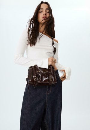 Giovane donna con lunghi capelli scuri, indossa una maglietta bianca a spalle scoperte, jeans larghi scuri e porta una piccola borsa a tracolla in pelle marrone scuro.