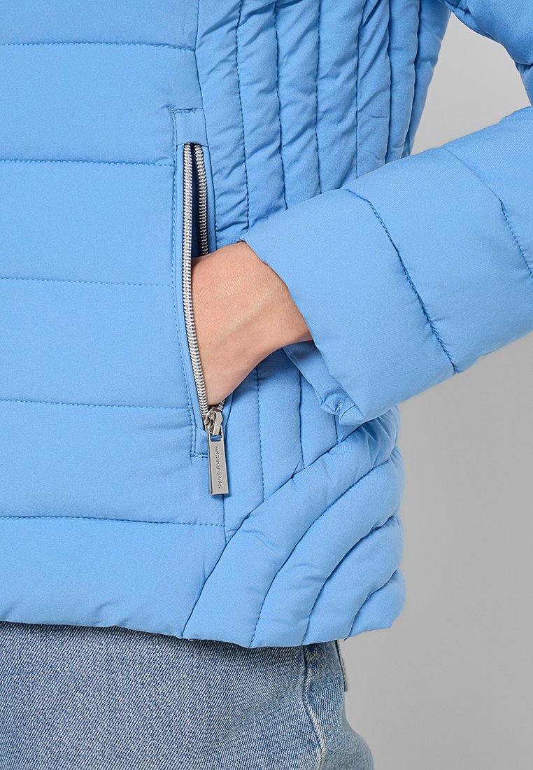Hellblaues Steppjacke mit gestepptem Design, ausgestattet mit einer seitlichen Reißverschlusstasche und einer weichen Textur. Die Ärmel sind eng anliegend für zusätzliche Wärme.