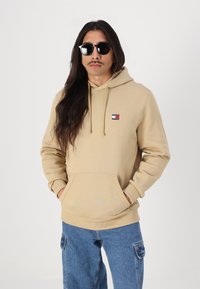 Beige hoodie with a front pocket and drawstring hood. Features a branded patch in red, white, and blue. Worn with blue cargo jeans.