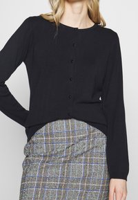 Schwarze Strickjacke mit langen Ärmeln und rundem Ausschnitt, mit schwarzen Knöpfen über die Vorderseite. Kombiniert mit einem karierter Rock.